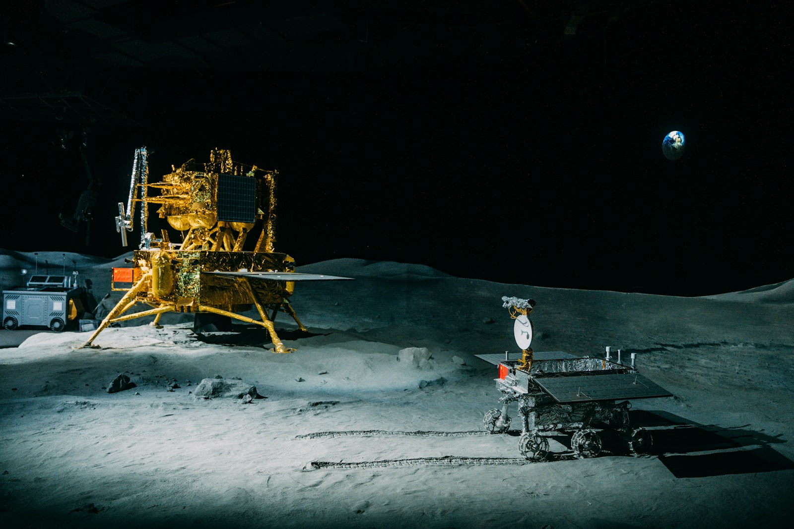 A lander and a small rover on the lunar surface, Earth visible in the black sky.